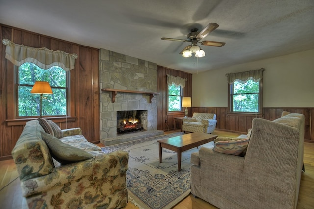 living room featuring a stone fireplace, wood walls, light wood-type flooring, a textured ceiling, and ceiling fan