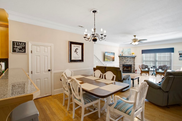 dining area with visible vents, ornamental molding, a stone fireplace, ceiling fan with notable chandelier, and light wood-style flooring