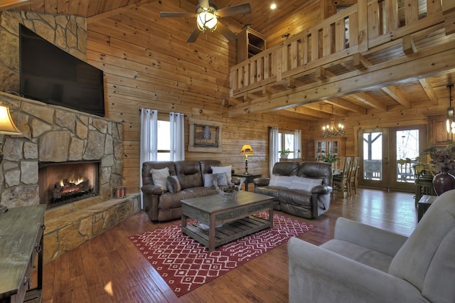 living area featuring a healthy amount of sunlight, wood walls, hardwood / wood-style floors, a stone fireplace, and ceiling fan with notable chandelier