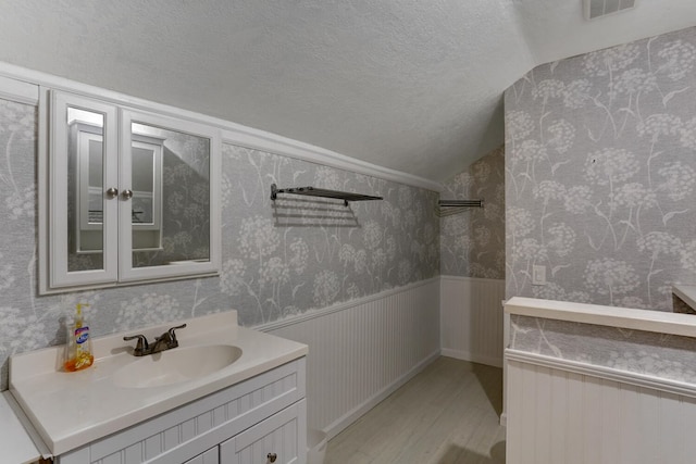 bathroom featuring wallpapered walls, a wainscoted wall, lofted ceiling, wood finished floors, and a textured ceiling