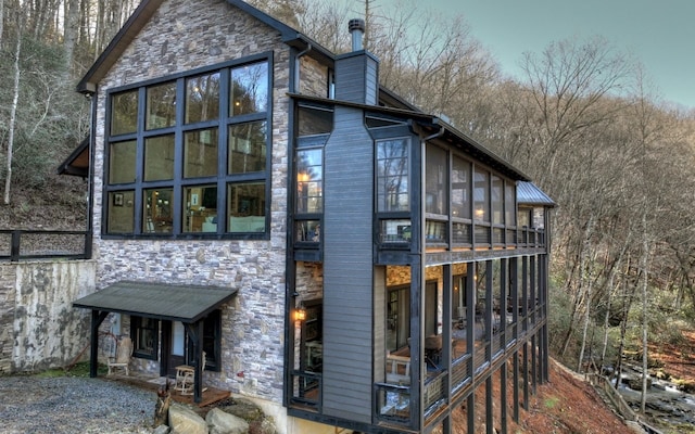 back of house featuring stone siding, a forest view, a chimney, and a sunroom
