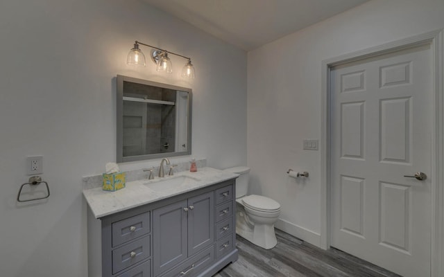 bathroom with toilet, vanity, wood finished floors, tiled shower, and baseboards