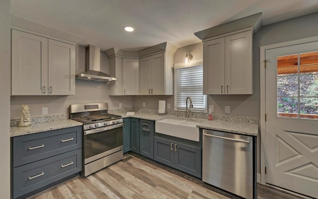 kitchen with light wood-style floors, light stone countertops, stainless steel appliances, wall chimney range hood, and a sink