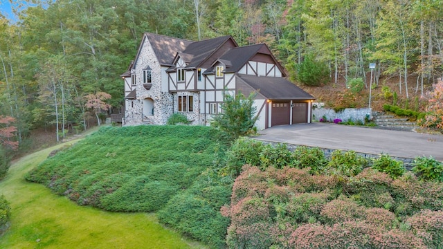 english style home with a forest view, driveway, a garage, stucco siding, and a front lawn