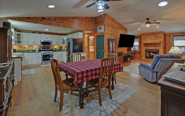 dining room featuring light wood-style floors, wood walls, a fireplace, ceiling fan, and vaulted ceiling