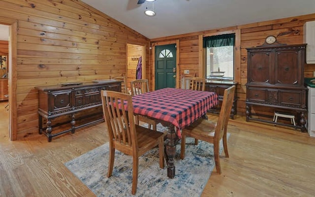 dining area featuring a ceiling fan, vaulted ceiling, wooden walls, and light wood-style floors