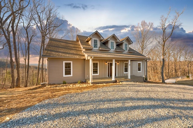 cape cod home featuring gravel driveway, covered porch, a shingled roof, and board and batten siding