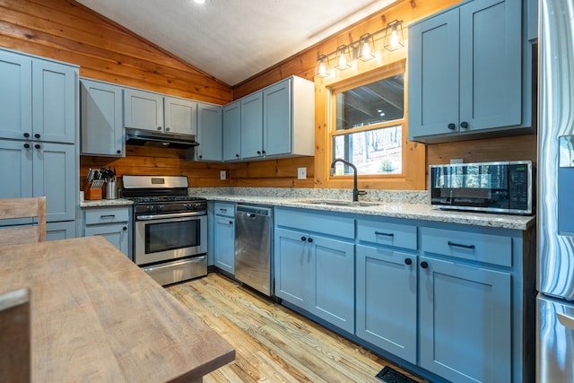 kitchen with appliances with stainless steel finishes, wooden walls, sink, lofted ceiling, and light hardwood / wood-style flooring