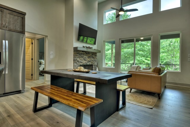 dining area featuring a ceiling fan, baseboards, light wood-style flooring, a stone fireplace, and a towering ceiling