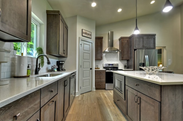 kitchen with pendant lighting, a sink, stainless steel appliances, wall chimney exhaust hood, and light wood finished floors