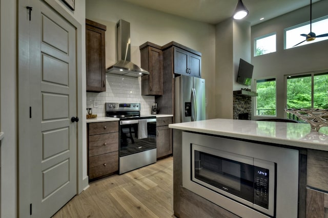 kitchen with light wood-style flooring, stainless steel appliances, light countertops, wall chimney range hood, and backsplash