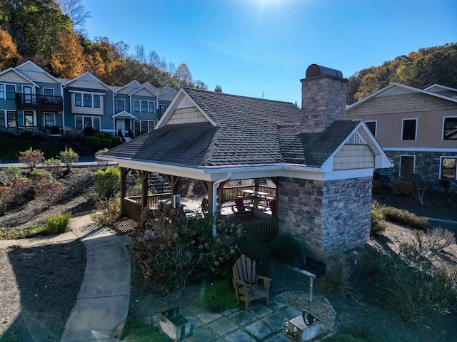 rear view of house featuring a gazebo, stone siding, roof with shingles, and a chimney