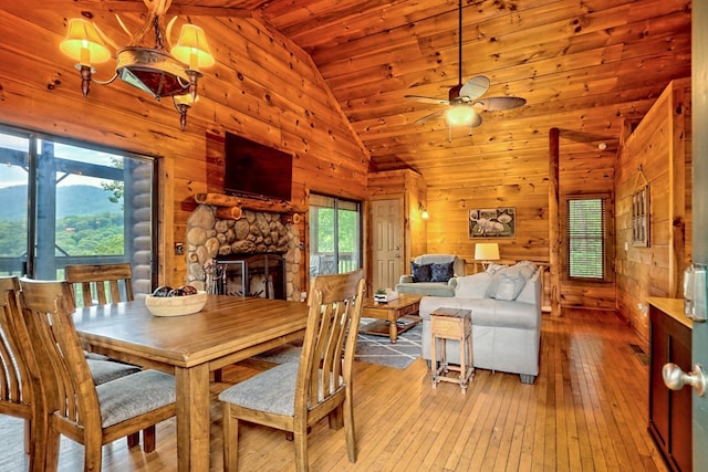 dining area with wooden walls, high vaulted ceiling, ceiling fan, a stone fireplace, and light wood-style floors