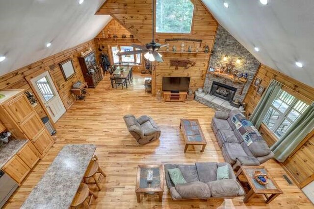 living room featuring ceiling fan, a stone fireplace, high vaulted ceiling, wood walls, and wood-type flooring