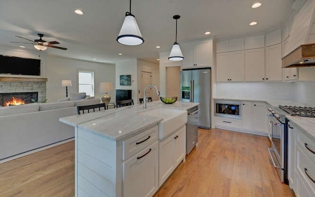 kitchen with white cabinetry, stainless steel appliances, a stone fireplace, an island with sink, and decorative light fixtures