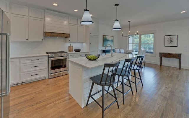 kitchen with premium range hood, decorative light fixtures, a center island with sink, white cabinetry, and stainless steel range with gas cooktop