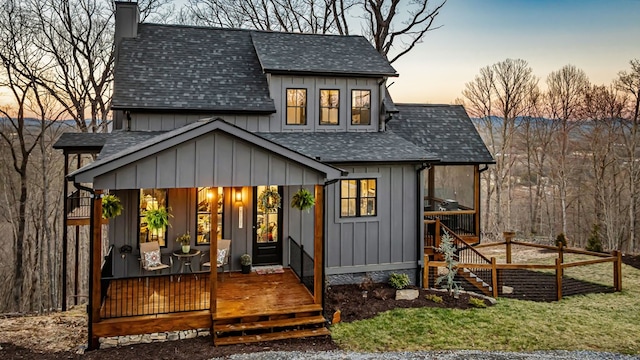 back of house at dusk featuring a porch, board and batten siding, roof with shingles, and a chimney