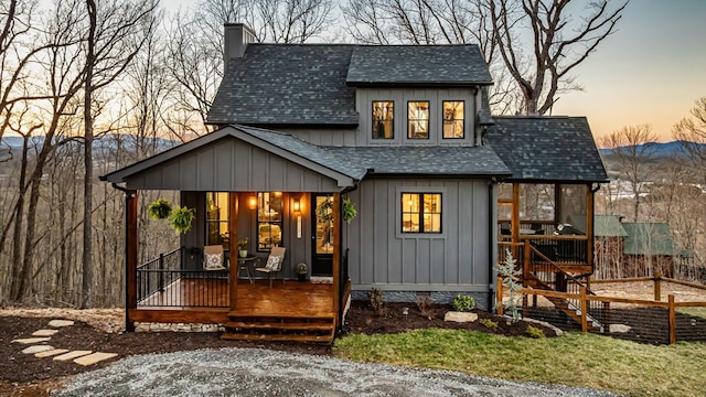 back of house at dusk featuring a shingled roof, covered porch, board and batten siding, and a chimney