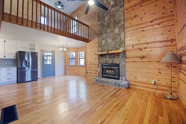 unfurnished living room with a stone fireplace, wood walls, light hardwood / wood-style flooring, a towering ceiling, and ceiling fan with notable chandelier