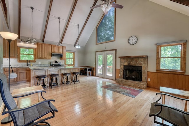 living area featuring high vaulted ceiling, wainscoting, light wood finished floors, ceiling fan, and a stone fireplace