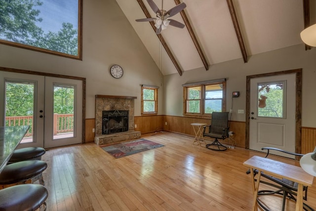 living area featuring high vaulted ceiling, wainscoting, light wood-style floors, a stone fireplace, and a ceiling fan