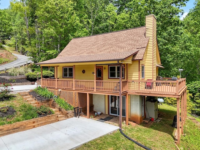 view of front of property featuring a shingled roof, a patio area, a wooden deck, a chimney, and a front lawn