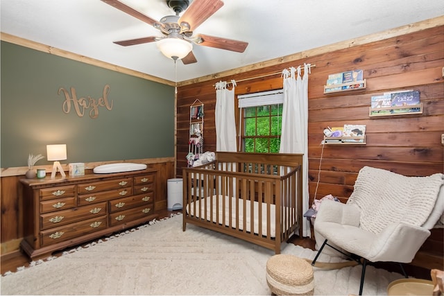 bedroom featuring a nursery area, ceiling fan, wooden walls, and wainscoting