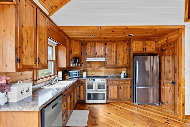 kitchen featuring under cabinet range hood, a sink, stainless steel appliances, wooden walls, and brown cabinetry