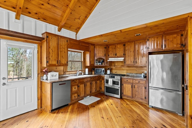 kitchen with brown cabinetry, vaulted ceiling with beams, a sink, stainless steel appliances, and under cabinet range hood