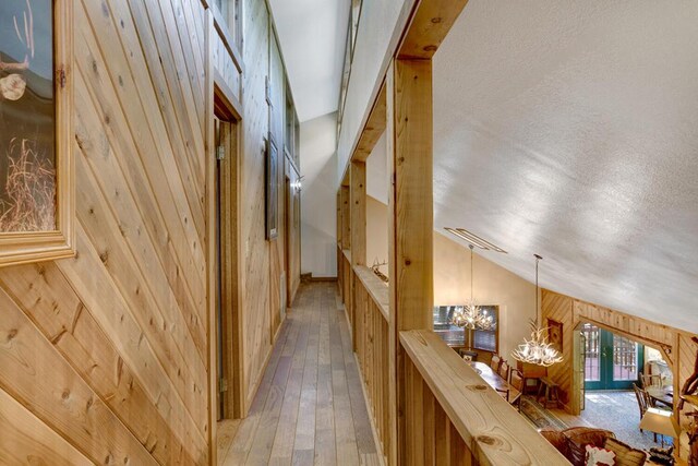 hallway featuring light hardwood / wood-style floors, a textured ceiling, lofted ceiling, wooden walls, and a notable chandelier