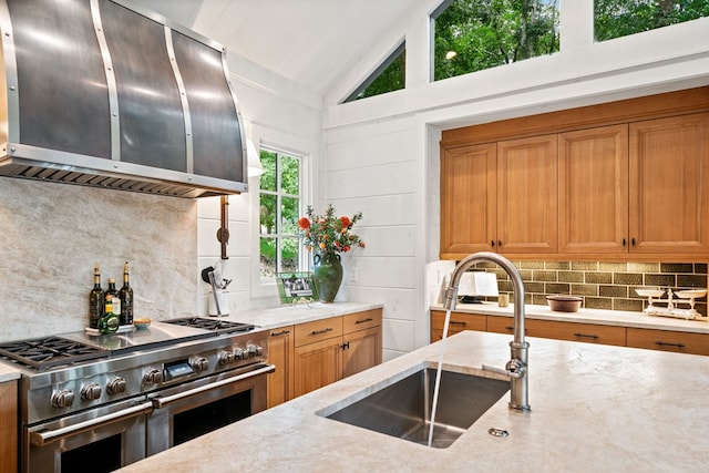 kitchen with ventilation hood, vaulted ceiling, light stone counters, range with two ovens, and a sink