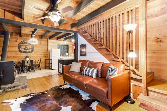 living room with wood walls, beamed ceiling, a wood stove, ceiling fan, and light hardwood / wood-style floors