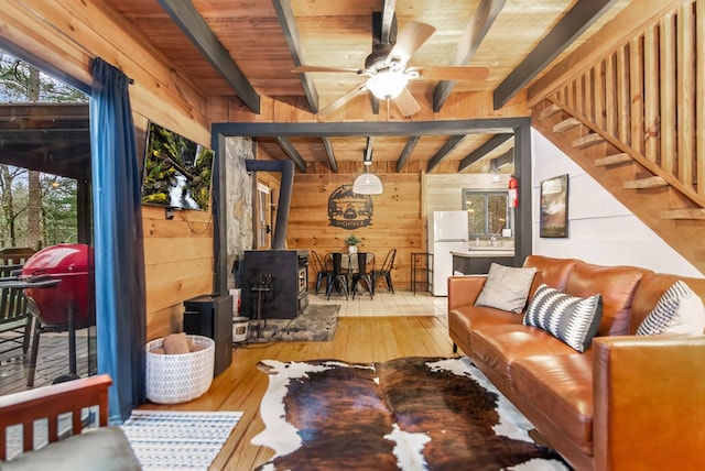 living room featuring ceiling fan, wooden walls, wood-type flooring, beamed ceiling, and a wood stove