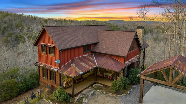 back of house at dusk with board and batten siding, a forest view, a chimney, and roof with shingles