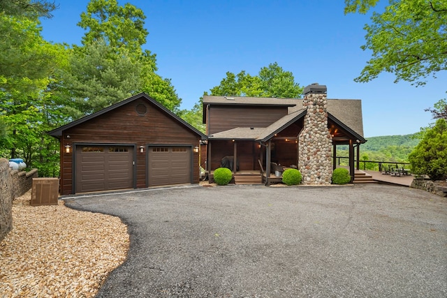 rustic home featuring covered porch, roof with shingles, aphalt driveway, a garage, and a chimney