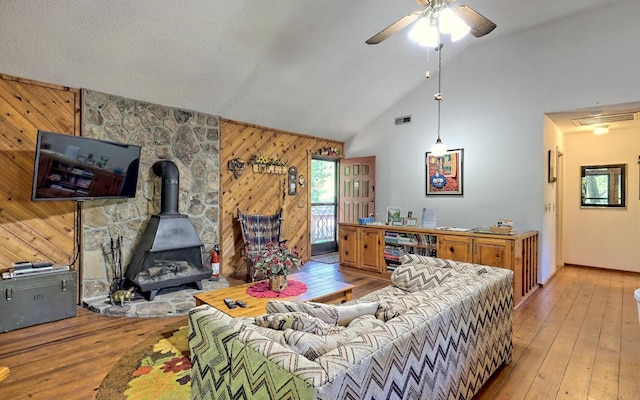 living room with a wood stove, wooden walls, vaulted ceiling, ceiling fan, and light hardwood / wood-style floors