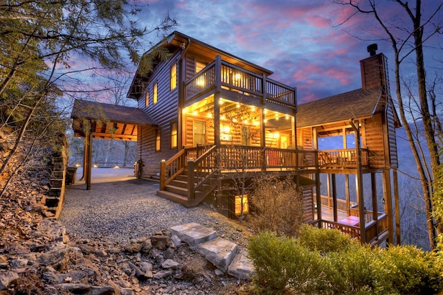 back of property at dusk with a chimney, faux log siding, roof with shingles, and a balcony