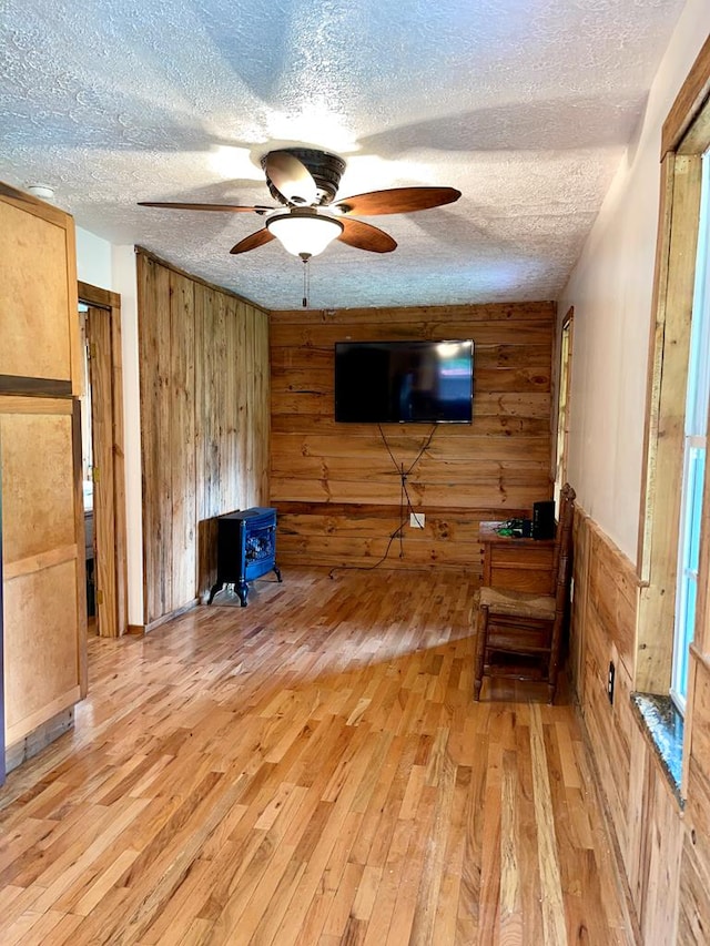 unfurnished living room with a wood stove, ceiling fan, light wood-type flooring, wood walls, and a textured ceiling