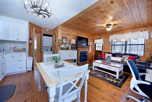 dining area with ceiling fan with notable chandelier, wood-type flooring, wooden ceiling, stacked washer and clothes dryer, and wood walls