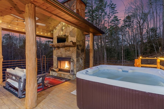 patio terrace at dusk with a wooden deck, a hot tub, and an outdoor stone fireplace