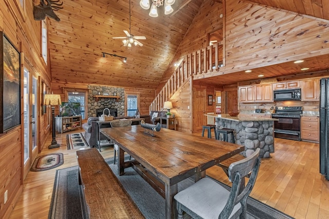 dining room featuring wooden walls, a ceiling fan, a stone fireplace, stairs, and light wood-type flooring