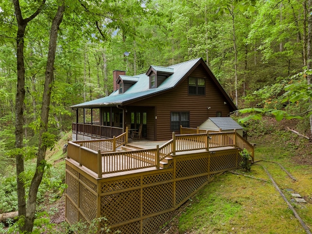 rear view of house with a view of trees, a wooden deck, a chimney, and metal roof