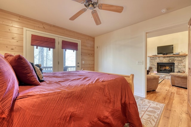 bedroom with access to exterior, light wood-style flooring, ceiling fan, a stone fireplace, and wooden walls