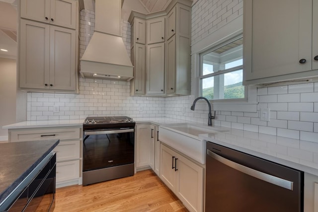kitchen featuring a sink, tasteful backsplash, custom exhaust hood, light wood-style flooring, and appliances with stainless steel finishes