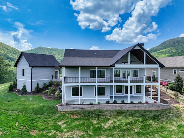 back of property with a lawn, a balcony, a mountain view, a patio area, and roof with shingles