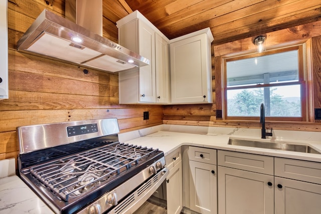 kitchen with sink, wall chimney range hood, stainless steel gas range, light stone counters, and wooden walls