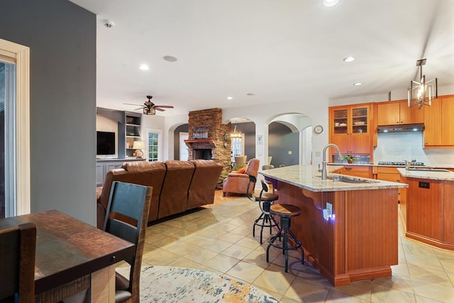 kitchen featuring light tile patterned flooring, sink, hanging light fixtures, a stone fireplace, and a kitchen island with sink