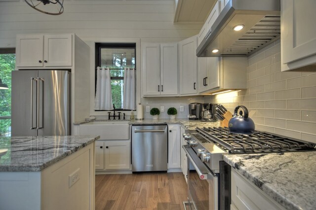 kitchen featuring exhaust hood, light stone countertops, light wood-type flooring, white cabinets, and premium appliances