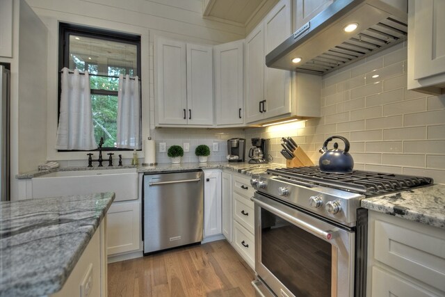 kitchen with range hood, appliances with stainless steel finishes, light hardwood / wood-style floors, light stone counters, and white cabinets