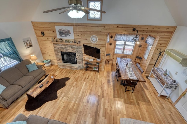 living room with a high ceiling, ceiling fan, wooden walls, a stone fireplace, and light wood-type flooring